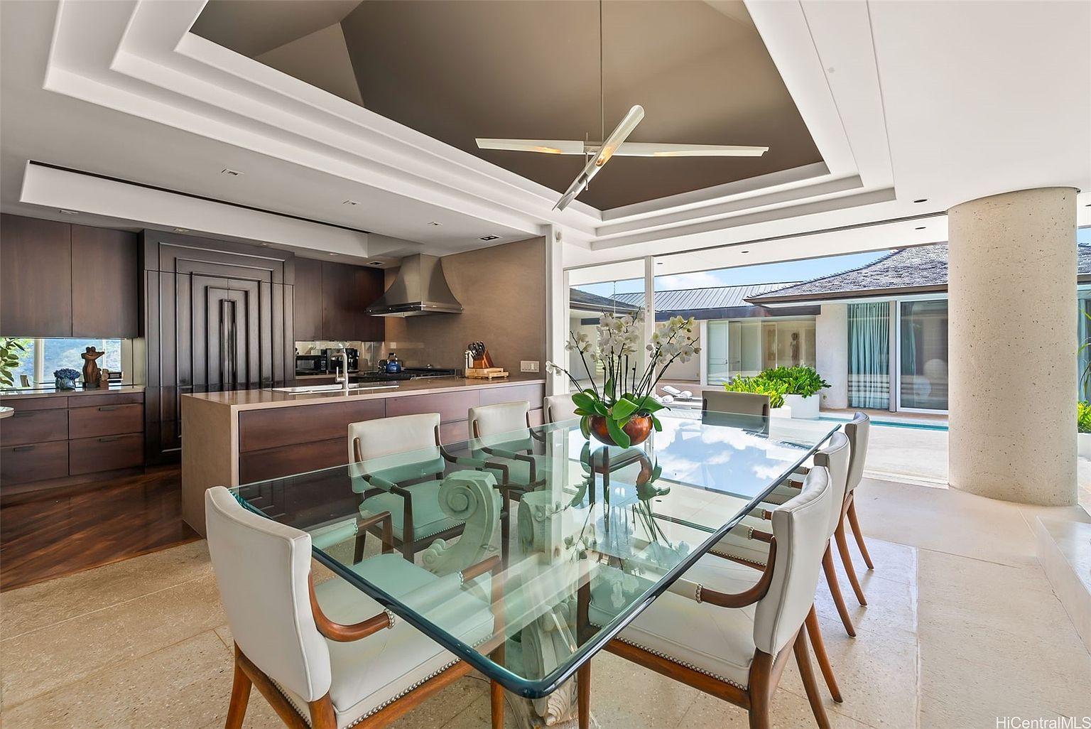 This interior shot showcases a modern dining area adjacent to the kitchen and with views of the exterior pool area. The focal point is a glass-topped dining table surrounded by white upholstered chairs with wooden frames. The space is well-lit, with a unique modern chandelier and natural light streaming in from the adjacent outdoor area, creating an inviting and luxurious atmosphere.