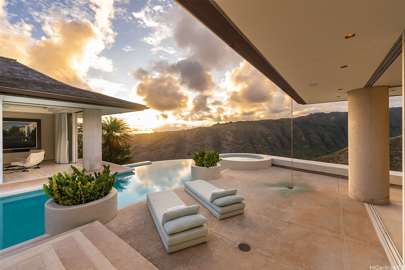 This image showcases a luxurious outdoor pool and spa area with stunning mountain views at sunset. The scene features two modern lounge chairs, lush potted plants, and a sleek infinity pool that blends seamlessly with the horizon. The architecture is contemporary, with clean lines and a neutral color palette, creating a serene and upscale ambiance.