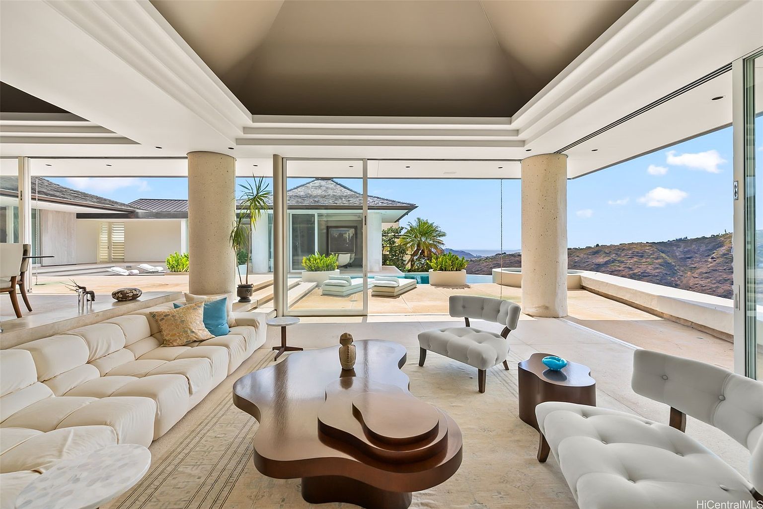 This is an interior shot of a luxurious living room that seamlessly blends indoor and outdoor spaces. The room features a large white sectional sofa, a unique wooden coffee table, and stylish armchairs, all set on a neutral-toned rug. Floor-to-ceiling glass doors open to a patio with views of a pool and the surrounding landscape, creating an open and airy atmosphere.