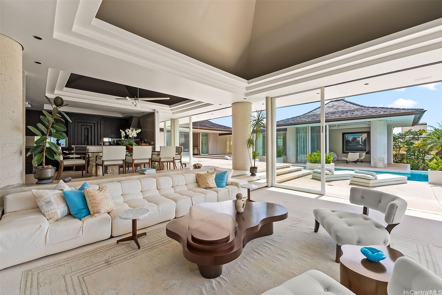This is a spacious and luxurious living room with an open floor plan that seamlessly connects to the outdoor area. The room features a large, sectional sofa, a unique coffee table, and floor-to-ceiling windows that offer stunning views of the pool and surrounding landscape. The design incorporates modern elements with a focus on natural light and indoor-outdoor living.