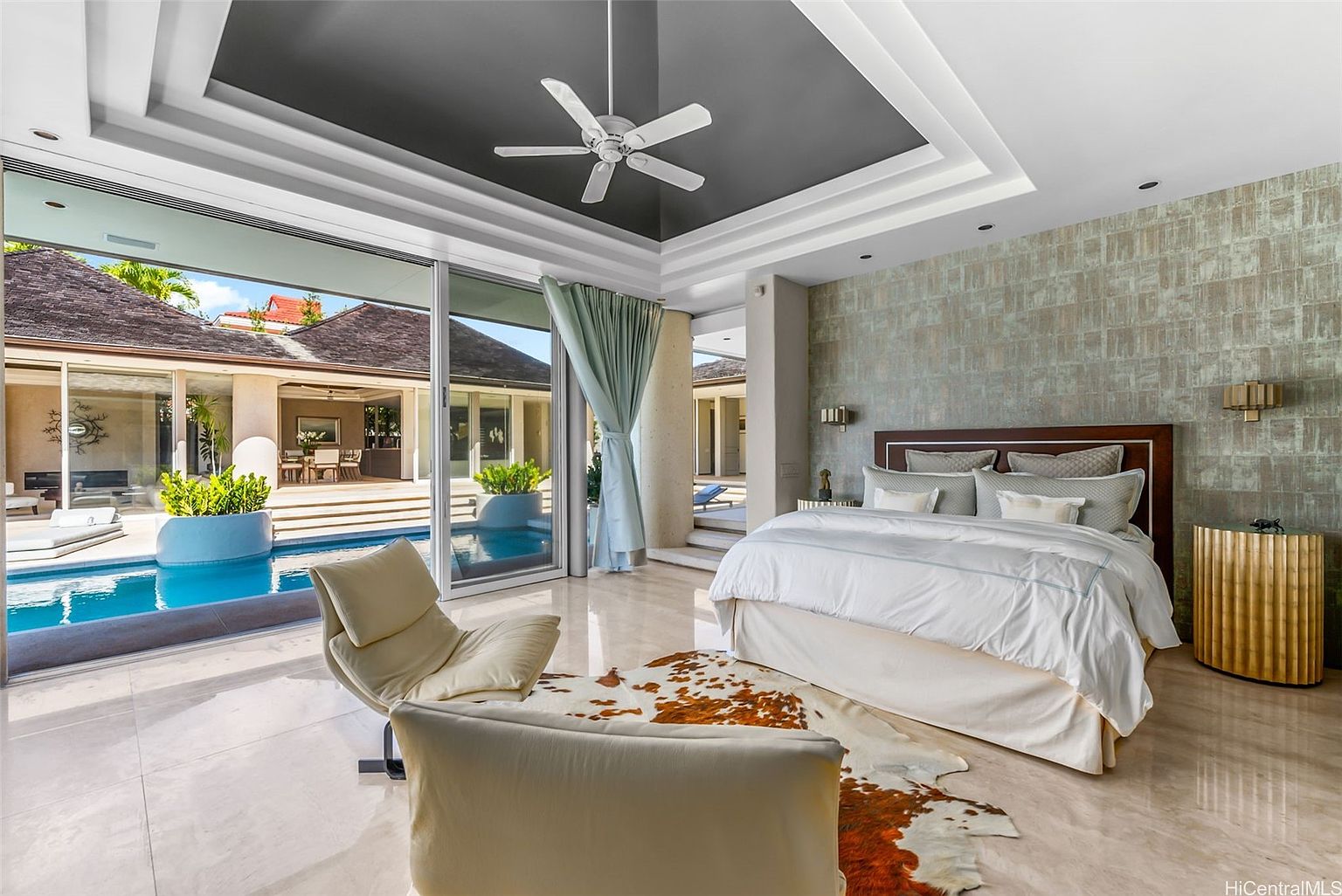 This is a luxurious primary bedroom with direct access to a pool area. The room features a large bed with a decorative headboard, a unique area rug, and stylish accent furniture. Sliding glass doors provide a seamless transition to the outdoor pool, enhancing the room's appeal and creating a resort-like atmosphere.
