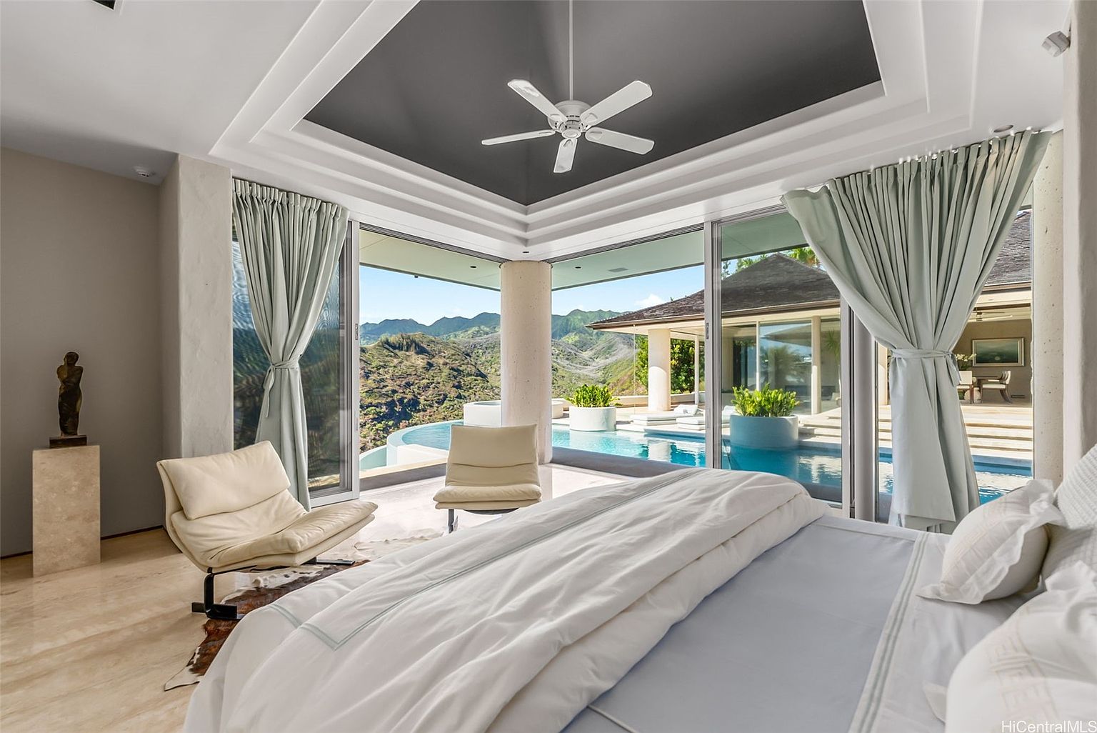This is a luxurious primary bedroom with floor-to-ceiling windows offering stunning views of the mountains and a private pool. The room features a neutral color palette, modern furniture, and a ceiling fan. The overall impression is one of tranquility and high-end living.