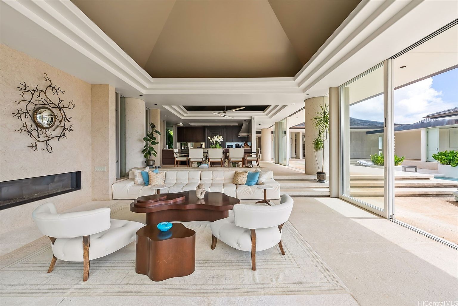 This is an interior shot of a luxurious living room featuring a modern fireplace, unique wall art, and stylish furniture including a white sofa and chairs. The open floor plan seamlessly connects to the dining area and outdoor patio through large glass doors, creating a bright and airy atmosphere. The neutral color palette and high ceilings contribute to the room's sophisticated and spacious feel.