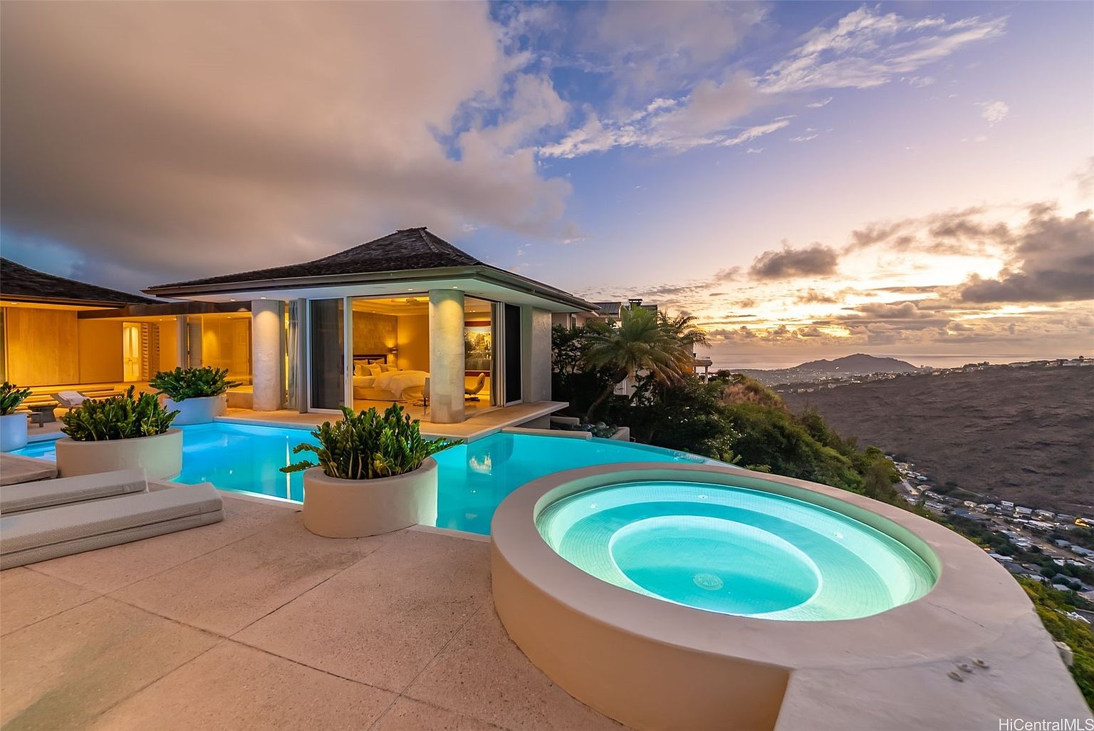 This image showcases a luxurious outdoor pool and spa area at sunset. The infinity pool seamlessly blends with the horizon, offering stunning views of the ocean and surrounding landscape. A modern architectural style is evident in the adjacent building, featuring large glass windows that open to the primary bedroom, and the circular spa adds a touch of elegance.