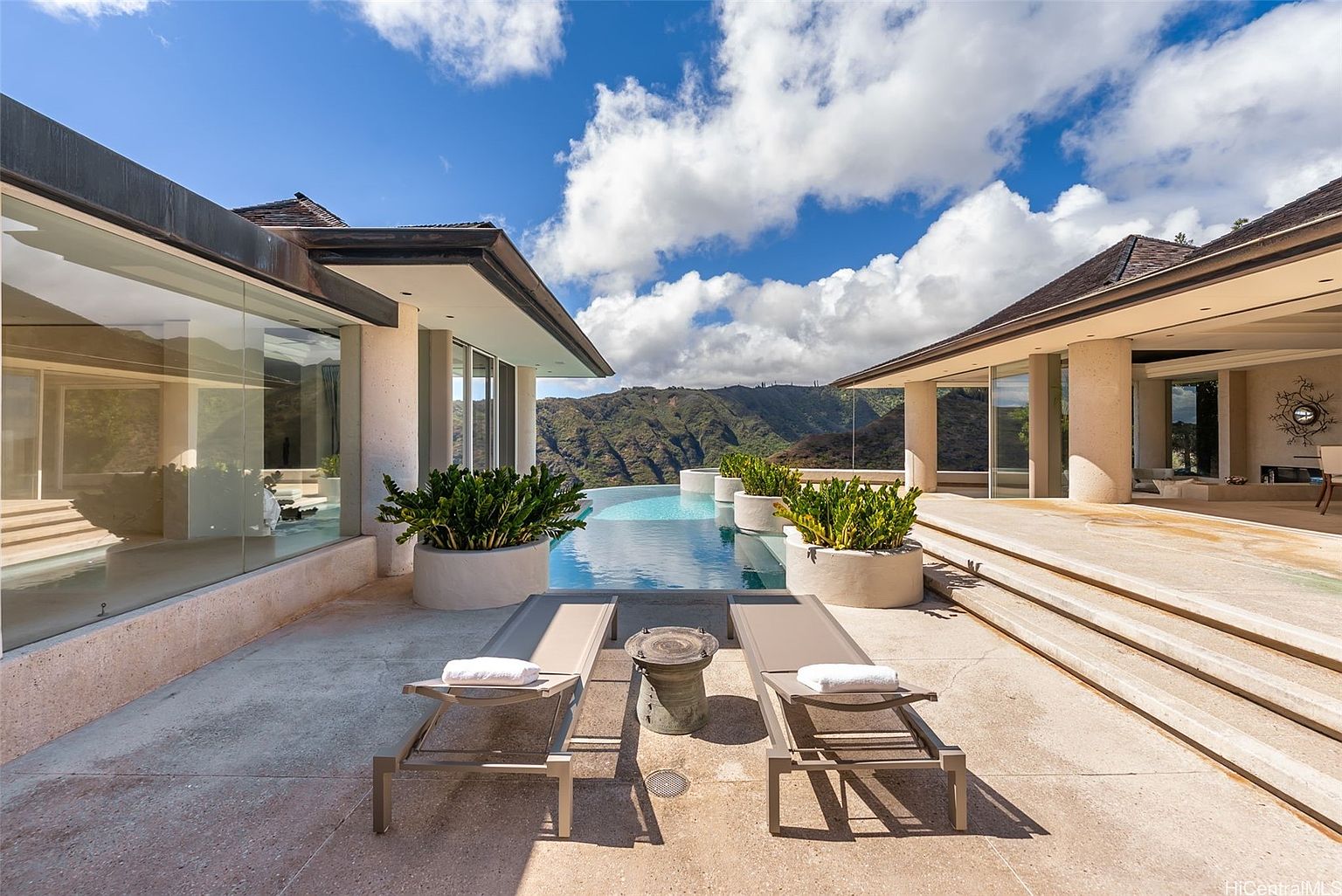 This image showcases a luxurious infinity pool area with stunning mountain views. Two lounge chairs flank a small table on the patio, inviting relaxation. The architecture features clean lines and large glass windows, blending indoor and outdoor spaces seamlessly, creating a serene and upscale ambiance.