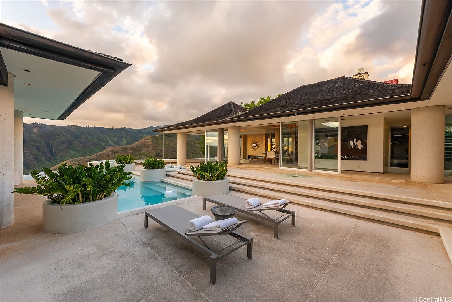 This image showcases a luxurious outdoor living space with a stunning pool and spa area. Two modern lounge chairs with plush towels are positioned on a spacious patio, leading to a contemporary home with expansive glass doors and architectural columns. The backdrop features a breathtaking mountain view, creating a serene and upscale ambiance.