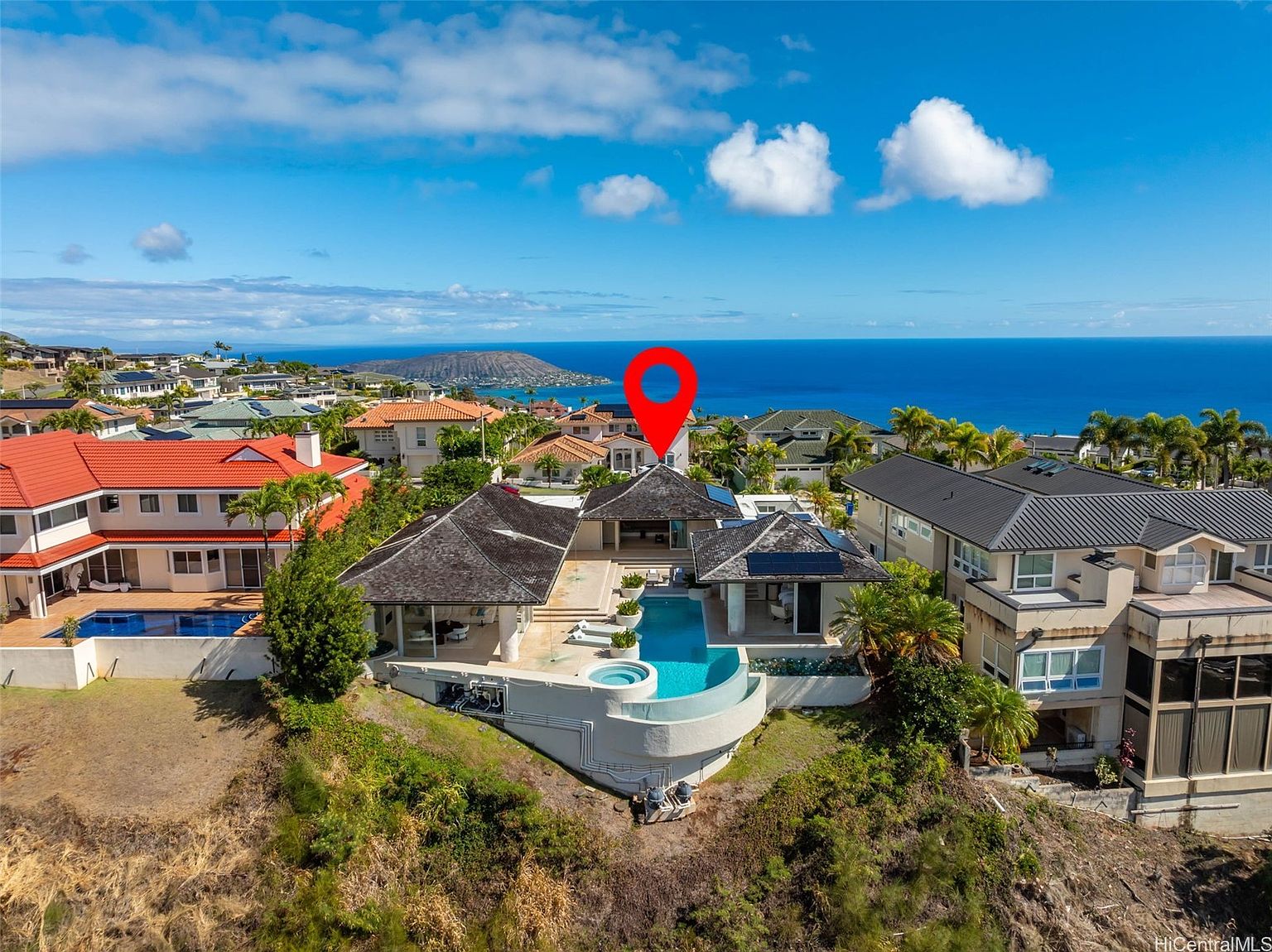 This aerial view showcases a luxurious home with a unique architectural design, featuring a dark-colored roof, a private pool and spa area, and well-maintained landscaping. The property is situated on a hillside, offering stunning ocean views and a sense of privacy. The surrounding neighborhood consists of similar upscale residences, creating an exclusive and desirable setting.