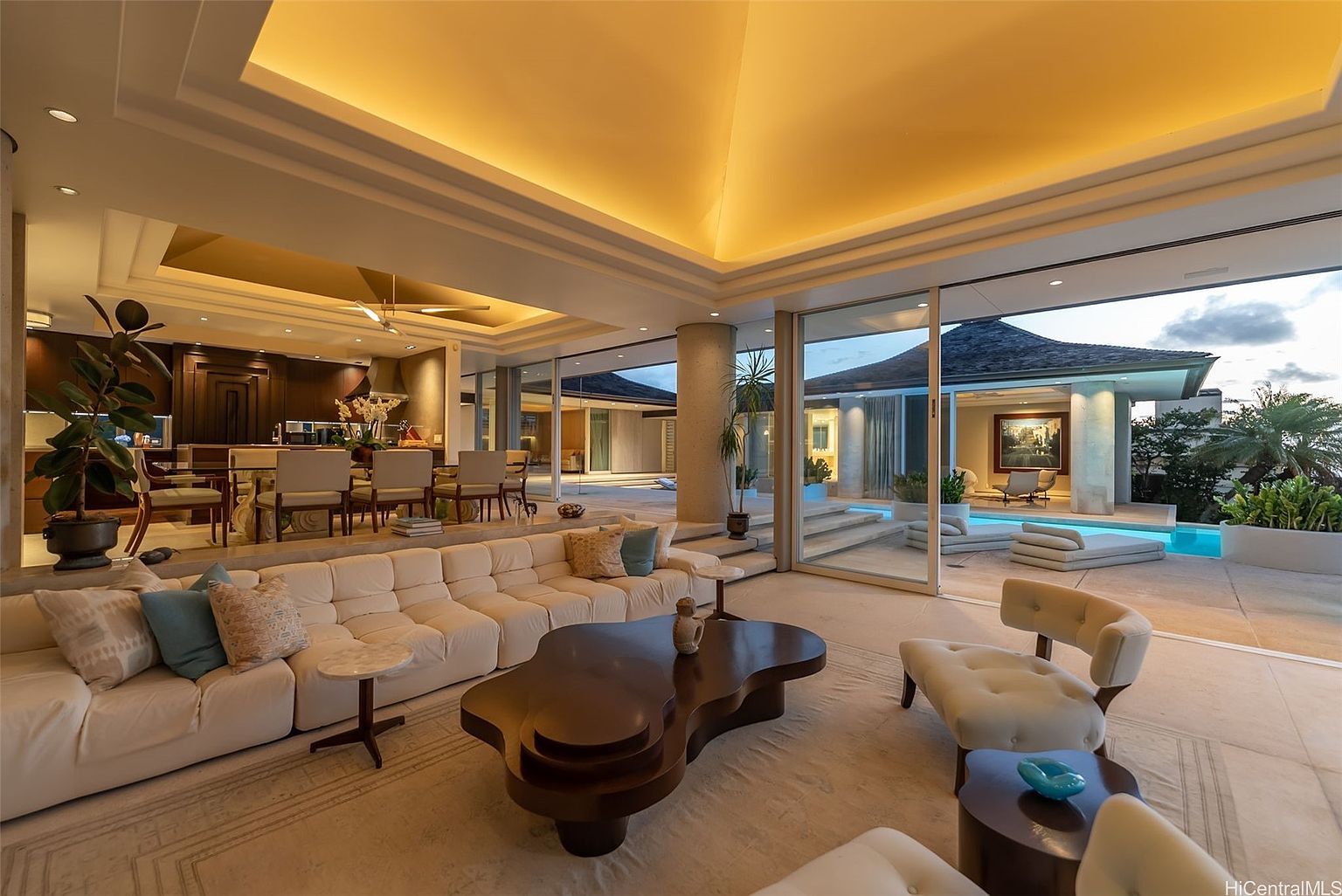 This is a luxurious living room that seamlessly blends indoor and outdoor living. The room features a large, comfortable white sectional sofa, a unique dark wood coffee table, and stylish armchairs. Floor-to-ceiling glass doors open to a patio with a pool, creating a bright and airy atmosphere with a clear view to the outside.