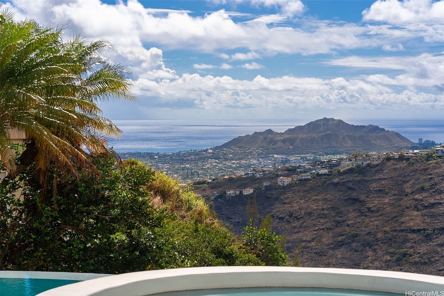This image showcases a stunning view from a property featuring a pool or spa. The foreground includes a portion of the pool's edge, leading to a lush, green landscape. In the distance, the ocean meets the horizon under a partly cloudy sky, with a prominent mountain visible, adding depth and character to the scene. The overall impression is one of luxury, tranquility, and breathtaking natural beauty.