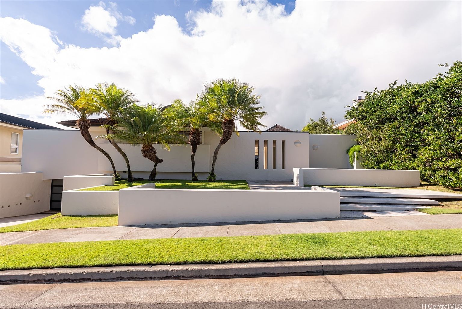 This is a front exterior view of a modern, single-story home featuring a white facade, minimalist design, and well-manicured landscaping. The property includes several palm trees, a low white wall bordering the lawn, and a multi-level entrance leading to the front door. The overall impression is clean, contemporary, and inviting.