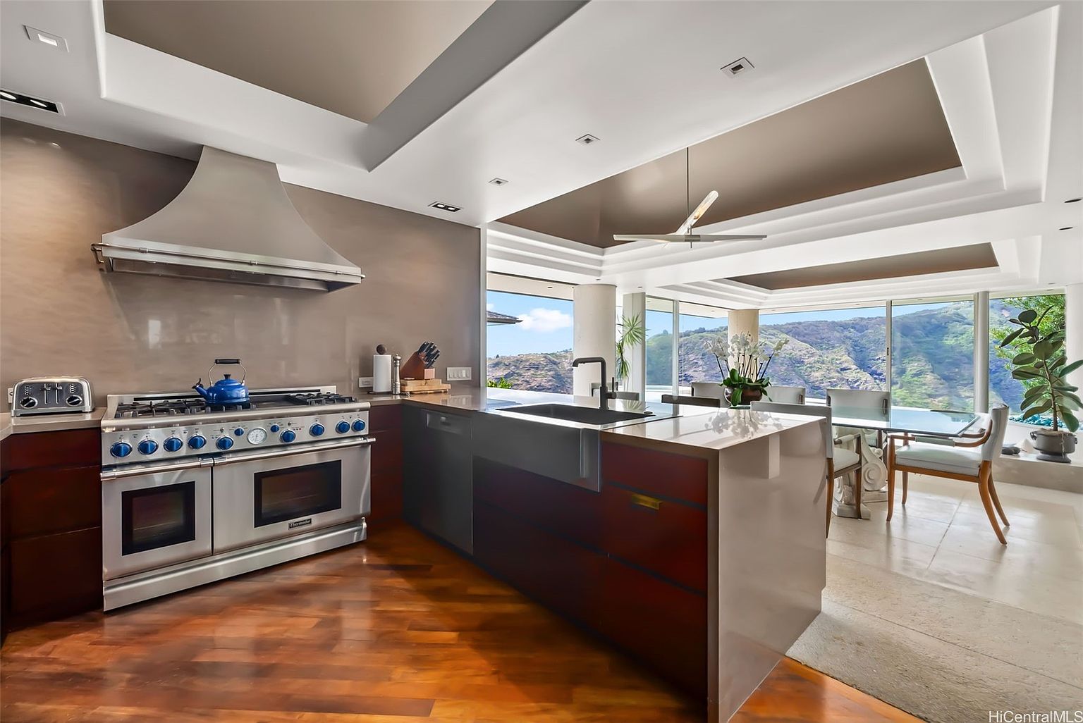 This is a well-lit, modern kitchen with a large island, stainless steel appliances, and dark wood cabinetry. The kitchen features a professional-grade range with a custom hood, a built-in sink, and an open layout that flows into the dining area with views of the landscape. The flooring is hardwood, and the overall impression is luxurious and functional.