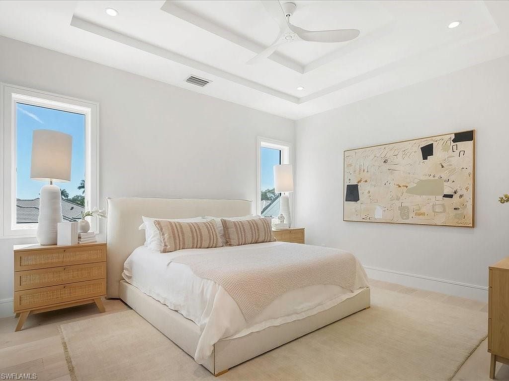 This is a bright and airy primary bedroom featuring a neutral color palette. The room includes a large bed with a padded headboard, two nightstands with lamps, and a piece of modern art on the wall. Natural light floods the space through two windows, creating a serene and inviting atmosphere.
