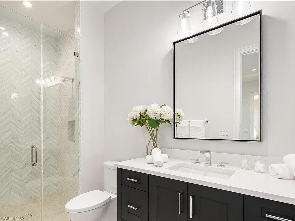 This is a well-lit bathroom featuring a glass-enclosed shower with herringbone tile, a toilet, and a vanity with a white countertop and dark cabinetry. A large mirror hangs above the sink, reflecting the clean, modern design. The space is decorated with white flowers and rolled towels, creating a fresh and inviting atmosphere.