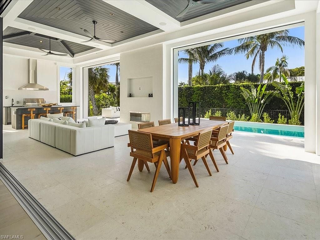 This is a bright and airy living space that seamlessly blends indoor and outdoor living. The open floor plan includes a modern kitchen, a comfortable seating area with a white sofa, and a dining area with a wooden table and woven chairs. Large sliding glass doors open to a patio and pool area, creating a luxurious and inviting atmosphere.