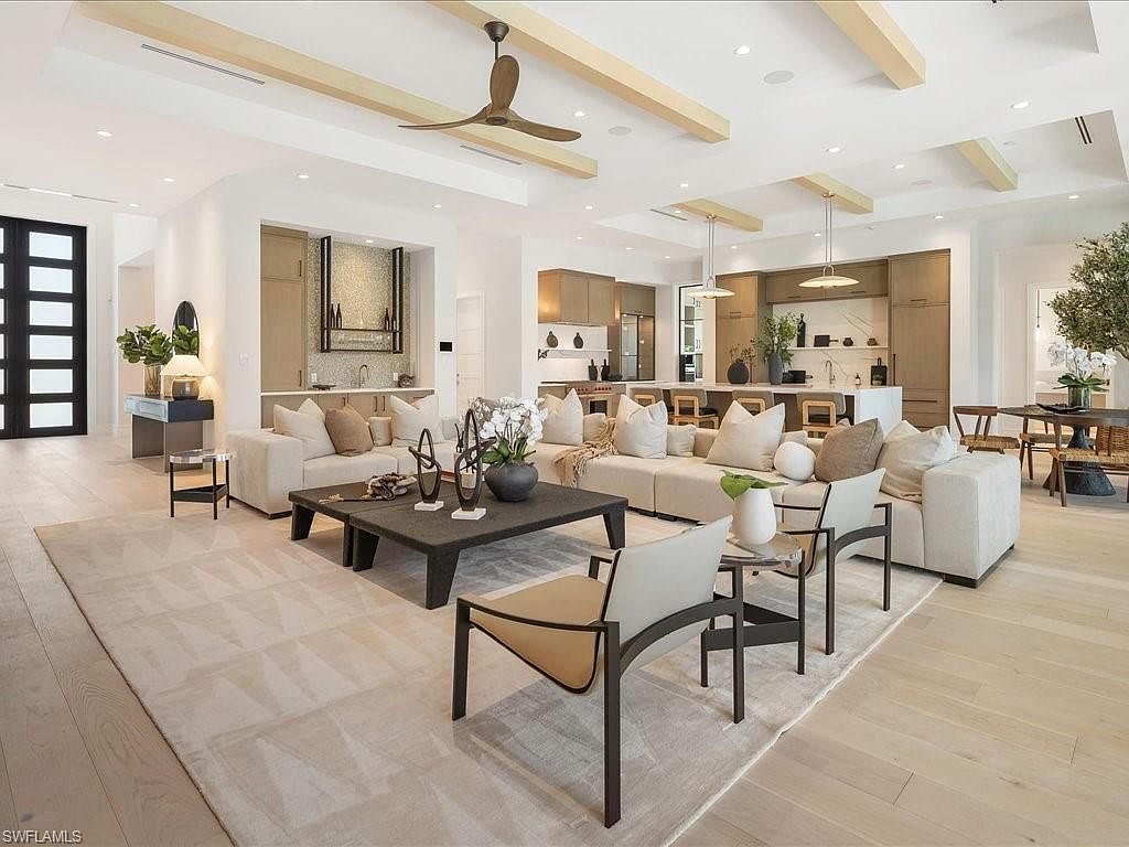 This is a spacious and modern living room featuring a large sectional sofa with neutral-toned cushions, a dark-colored coffee table with decorative objects, and two armchairs. The room has high ceilings with wooden beams and recessed lighting, and it opens to the kitchen area. The flooring is light-colored wood, and a large area rug defines the seating area, creating a warm and inviting atmosphere.