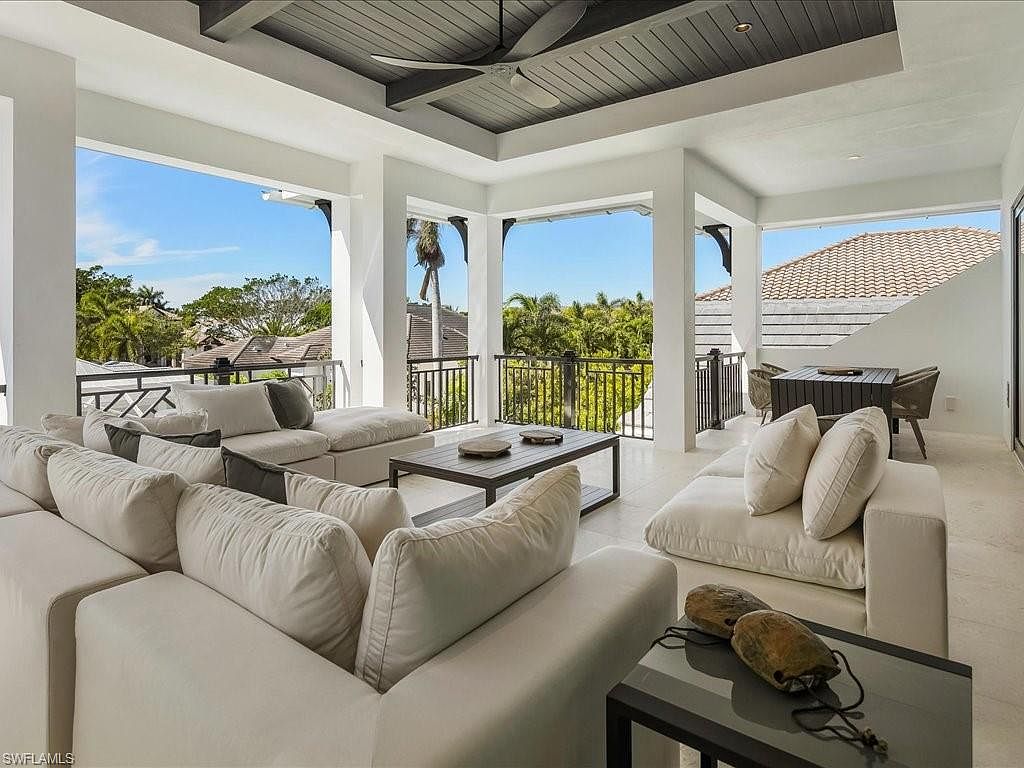 This image showcases a luxurious outdoor living space, featuring comfortable white sofas and a coffee table, perfect for relaxation and entertaining. The space is partially covered with a dark gray ceiling and supported by white pillars, offering views of the surrounding lush greenery and rooftops. The overall impression is one of sophisticated comfort and seamless indoor-outdoor living.