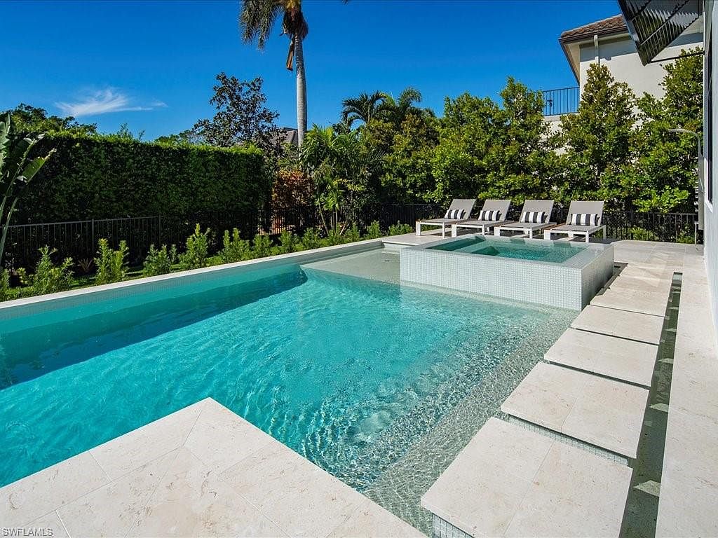 This image showcases a luxurious backyard pool and spa area. The pool features clear turquoise water and is surrounded by light-colored stone tiling. A raised spa with mosaic tiling is integrated into the pool design, and lounge chairs are arranged on the deck, creating an inviting and relaxing outdoor space.