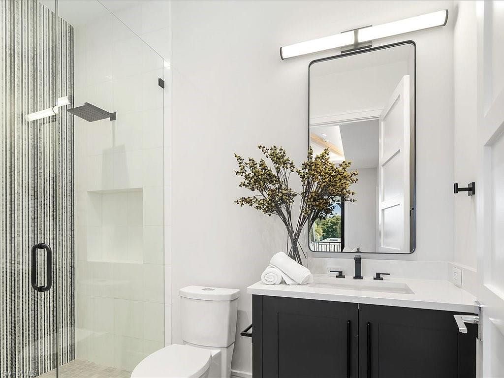 This is a well-lit bathroom featuring a modern design. The space includes a glass-enclosed shower with vertically striped tile, a white toilet, and a vanity with a dark cabinet and white countertop. A rectangular mirror hangs above the sink, complemented by a modern light fixture, and a vase with decorative branches adds a touch of elegance.