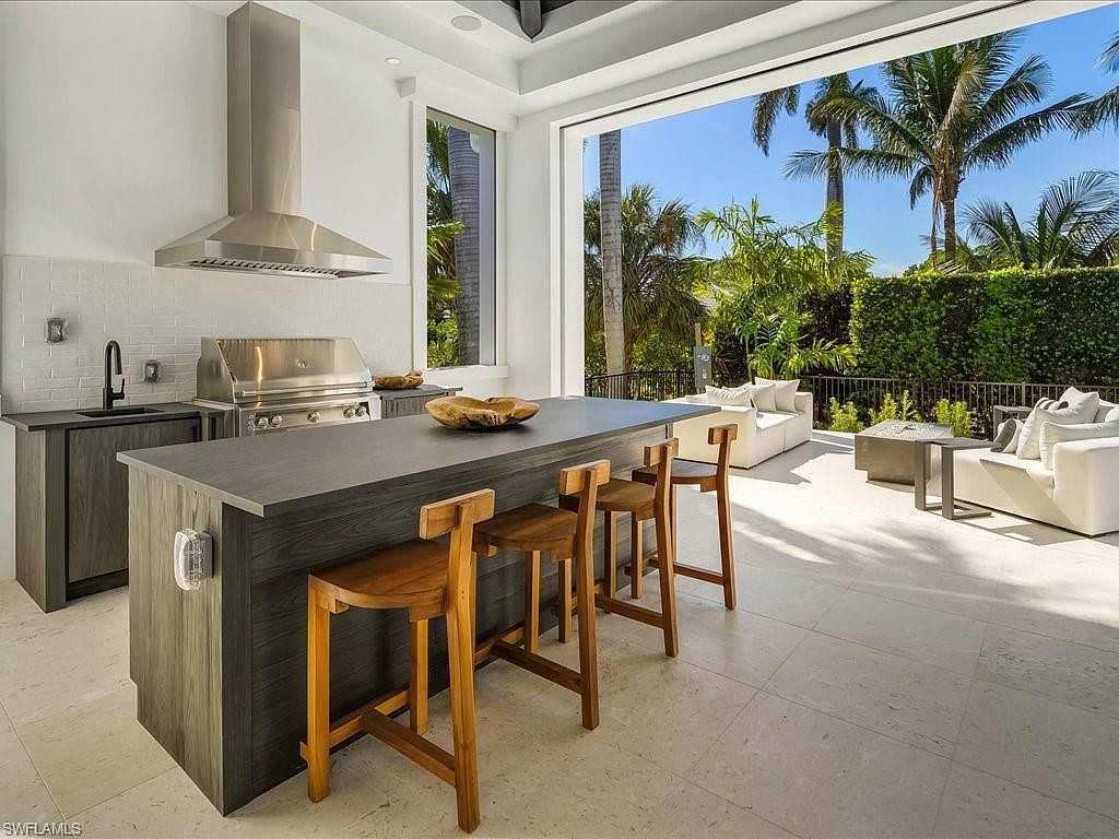 This image showcases an outdoor kitchen area seamlessly blending indoor and outdoor living. The kitchen features a built-in grill, sleek countertops, and bar seating, while the open layout leads to a patio with comfortable seating and lush landscaping. The design emphasizes modern luxury and effortless entertaining.
