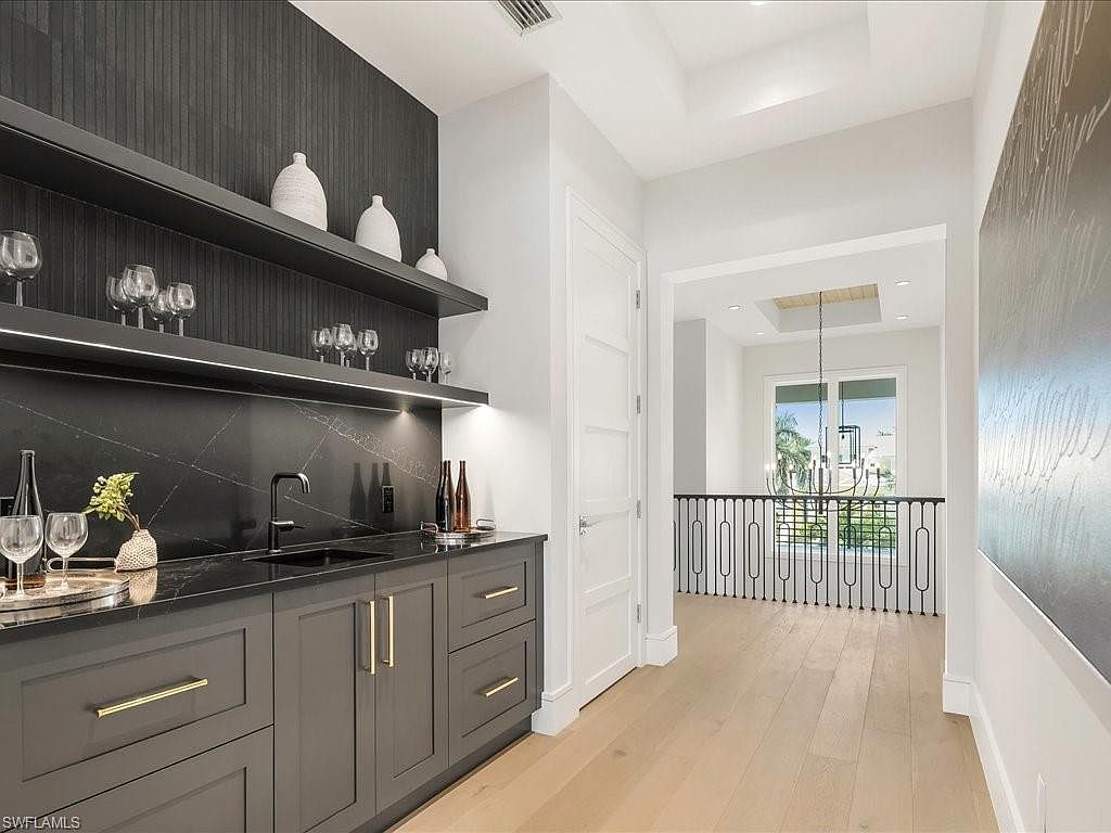 This interior shot showcases a modern hallway featuring a stylish wet bar area with dark cabinetry, gold hardware, and open shelving displaying glassware and decorative items. The hallway leads to a bright, open space with a view of the outdoors through a large window, complemented by light wood flooring and white walls, creating a clean and sophisticated aesthetic. The perspective is from a medium distance, allowing for a comprehensive view of the hallway and its features.