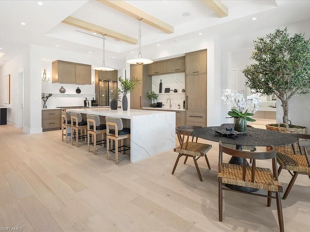 This is a bright and spacious kitchen featuring a large waterfall island with bar seating, custom cabinetry in a light wood tone, and modern pendant lighting. The open floor plan includes a dining area with a round table and unique chairs, complemented by a large potted tree, creating an inviting and stylish atmosphere. The perspective is a wide shot, showcasing the kitchen's layout and design elements.