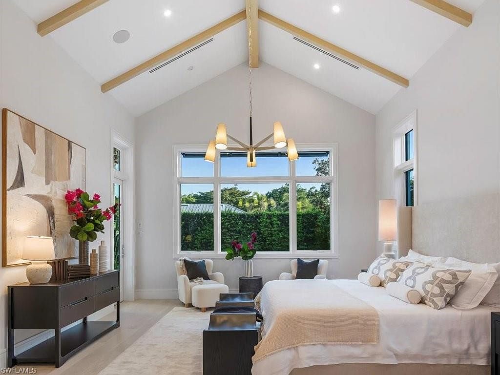This is a bright and airy primary bedroom featuring a high vaulted ceiling with exposed wooden beams and a large window offering a view of lush greenery. The room is decorated in a neutral color palette with a large abstract painting, modern chandelier, and stylish furniture, creating a serene and luxurious atmosphere. The perspective is from the foot of the bed, capturing the entire room.