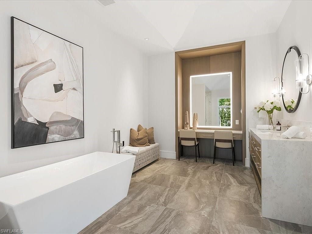 This is a luxurious primary bathroom featuring a freestanding white bathtub positioned beneath a large abstract art piece. The room includes a vanity area with a lighted mirror and seating, as well as a marble countertop with dual sinks and modern fixtures. The flooring is a large-format tile with a marble-like pattern, contributing to the bathroom's elegant and spa-like ambiance.