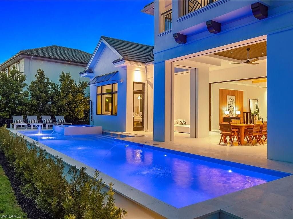 This image showcases a luxurious outdoor pool area at dusk, featuring a long, rectangular pool with integrated spa and water features. The pool is illuminated with blue lighting, creating a serene ambiance. The adjacent patio offers an open-air dining space, seamlessly blending indoor and outdoor living, while manicured landscaping adds to the property's appeal.