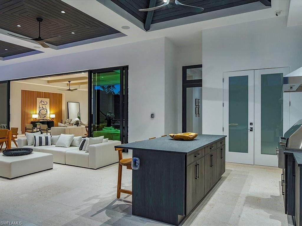 This interior shot showcases a modern living room that seamlessly blends with the kitchen area. The living room features a large white sectional sofa, a sleek ottoman, and a view into an adjacent room through wide sliding glass doors. The kitchen island is prominently displayed, adding to the open-concept feel of the space.