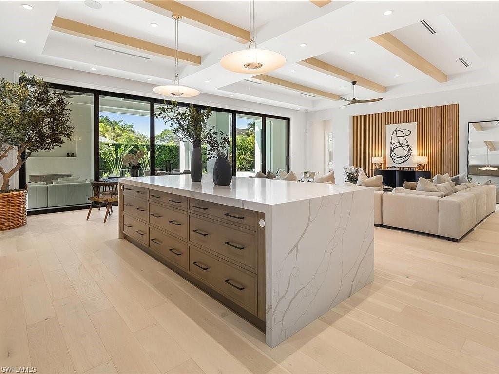This is a bright and spacious kitchen featuring a large island with a white countertop and wooden cabinetry. The open-concept design seamlessly connects the kitchen to the living area, enhancing the sense of space. Large windows provide ample natural light and views of the outdoors, while modern lighting fixtures illuminate the area.