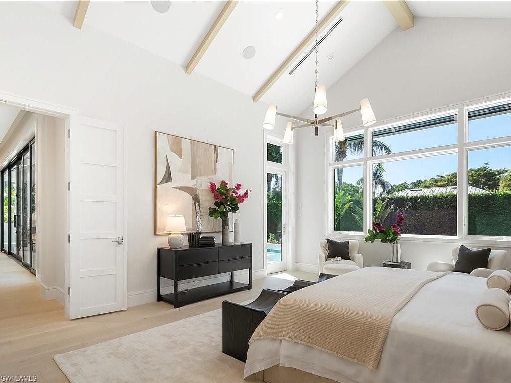 This is a bright and airy primary bedroom featuring a high vaulted ceiling with exposed beams and a large window offering natural light and views of the outdoors. The room is decorated in a neutral color palette with a large abstract painting, a dark wood dresser, and a comfortable bed with plush pillows and a throw blanket. The overall impression is one of luxury and relaxation.