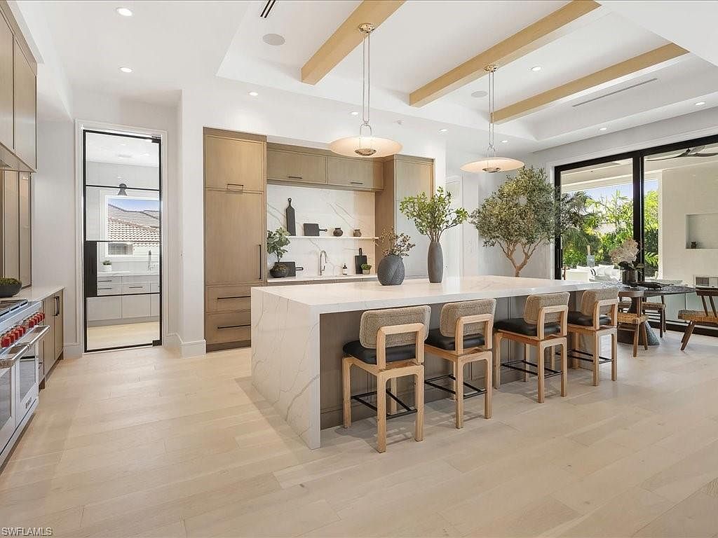 This is a bright and modern kitchen featuring a large island with seating, light wood flooring, and custom cabinetry. The kitchen is well-lit with pendant lighting and natural light from a large sliding glass door leading to the outside. The space is designed with a clean and contemporary aesthetic, perfect for entertaining and everyday living.