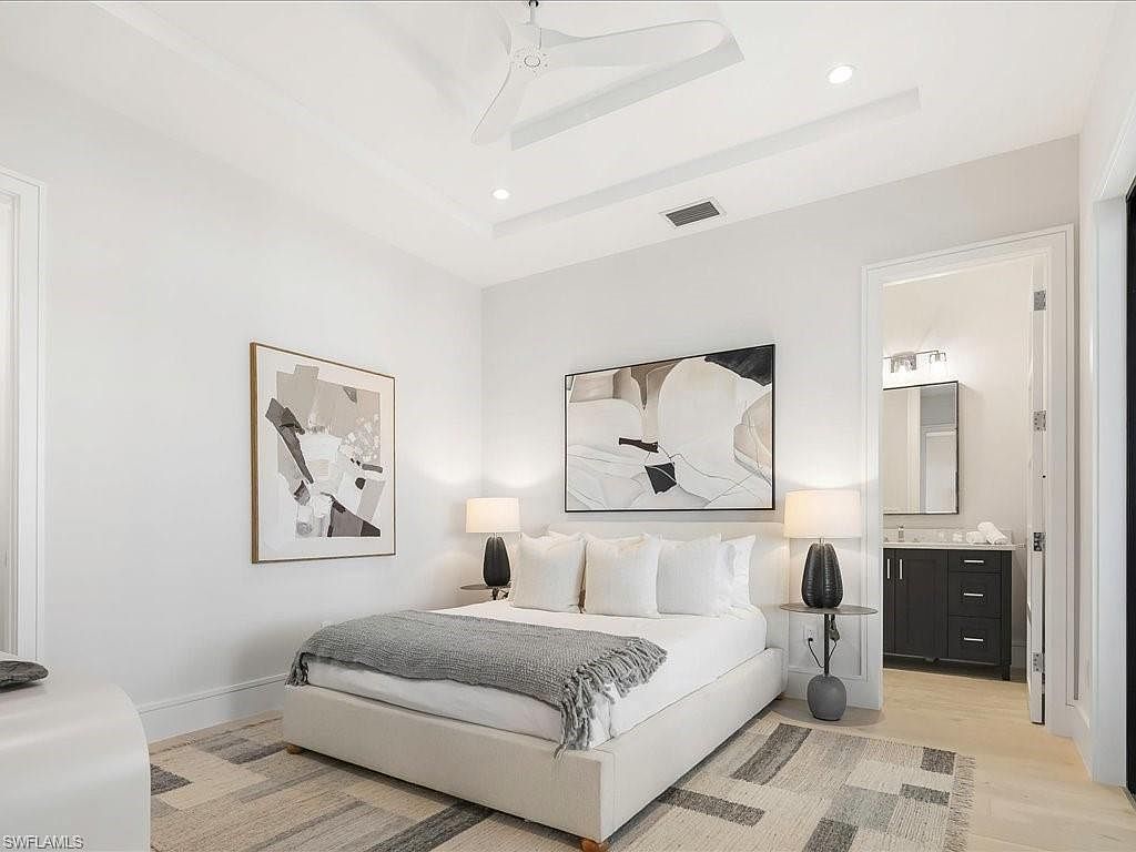 This is a bright and airy bedroom featuring a neutral color palette. The room includes a bed with white linens and a gray throw, flanked by side tables with lamps. Artwork adorns the walls, and a doorway leads to an adjacent bathroom, creating a serene and inviting space.