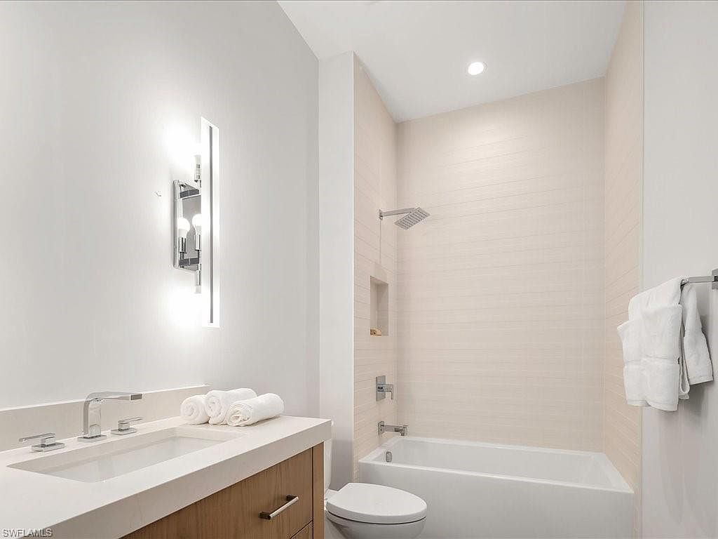 This is a well-lit bathroom featuring a modern vanity with a white countertop and light wood cabinetry. A contemporary chrome light fixture illuminates the space above the sink, where neatly folded white towels are placed. The bathroom also includes a bathtub and shower combination with light-colored tile, creating a clean and inviting atmosphere.