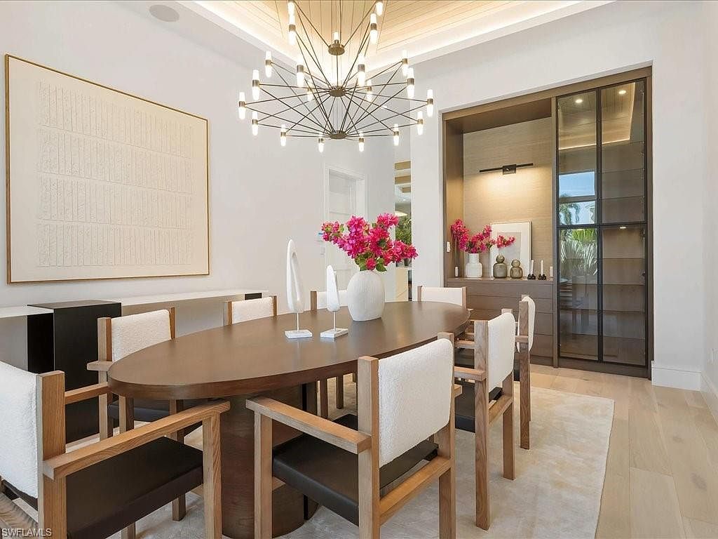 This is an interior shot of a dining room featuring an oval dining table with seating for eight. The room is decorated with a large abstract painting, a modern chandelier, and a built-in cabinet with glass doors. The overall style is elegant and contemporary, creating a sophisticated dining experience.