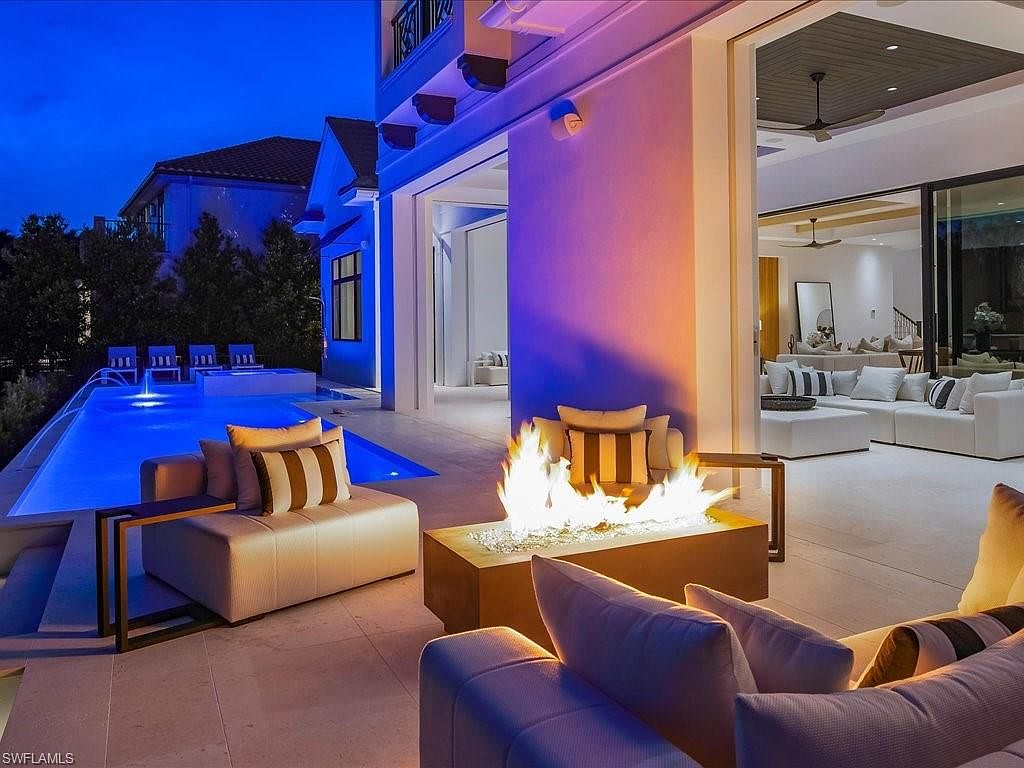 This image showcases a luxurious outdoor patio area at dusk, featuring a modern fire pit surrounded by comfortable lounge seating with striped pillows. The patio seamlessly connects to an open-concept interior through large sliding glass doors, offering a glimpse of the stylish living room. A sleek pool with illuminated water features adds to the upscale ambiance, creating an inviting space for relaxation and entertainment.
