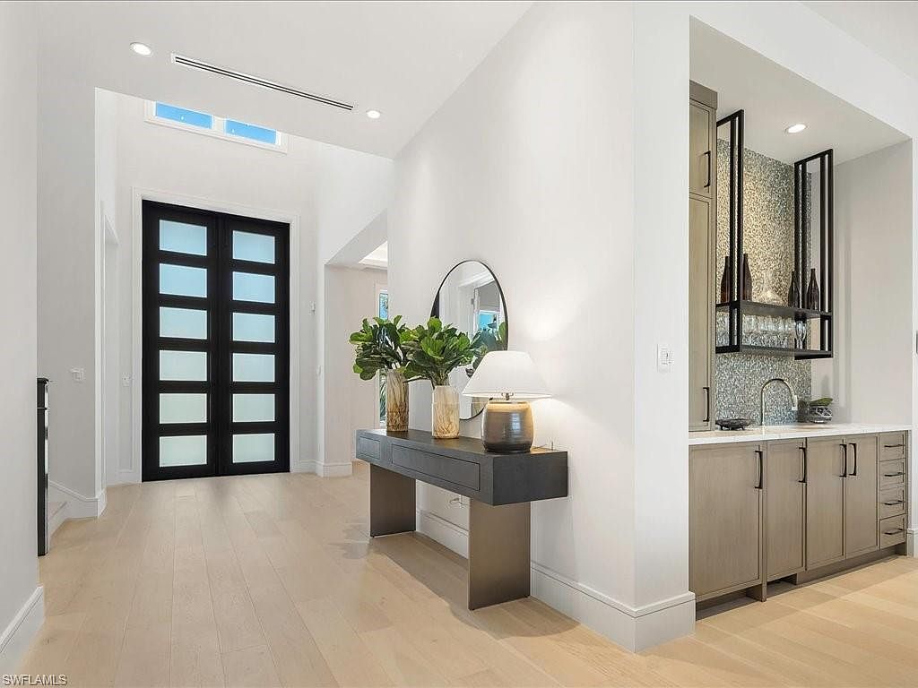 This interior shot showcases a bright and modern hallway with light wood flooring and white walls. A striking black double door with frosted glass panels serves as a focal point, complemented by a stylish console table with a round mirror and a table lamp. A built-in bar area with mosaic tile backsplash adds a touch of luxury and functionality to the space.