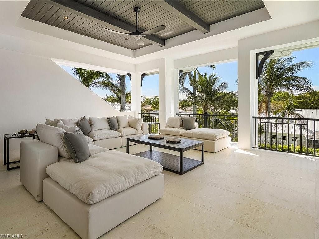 This is a bright and airy living room featuring a large, modular sectional sofa with plush cushions and neutral tones. The room opens to an outdoor balcony with palm tree views, enhancing the indoor-outdoor living experience. A dark-framed coffee table sits in the center, and the ceiling has a modern, dark-toned design with a ceiling fan.