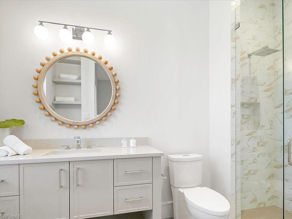 This is a bright and clean bathroom featuring a white vanity with a light countertop, a round mirror with a wooden beaded frame, and a modern toilet. The shower is enclosed with glass and has marble-like tiling. The overall impression is fresh and contemporary, perfect for a guest bathroom.