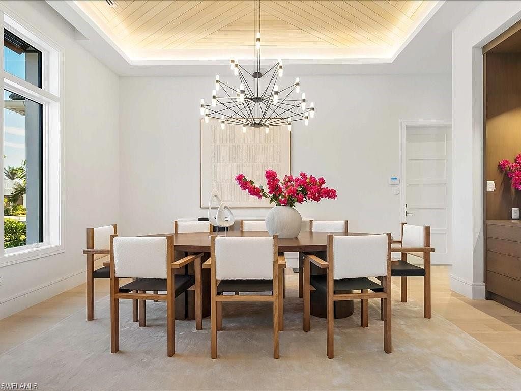 This is an interior shot of a dining room featuring a round wooden table surrounded by eight chairs with wooden frames and white cushions. A modern chandelier hangs above the table, and a large piece of artwork is displayed on the wall. The room is bright and airy, with a neutral color palette and hardwood flooring, creating an elegant and inviting atmosphere.