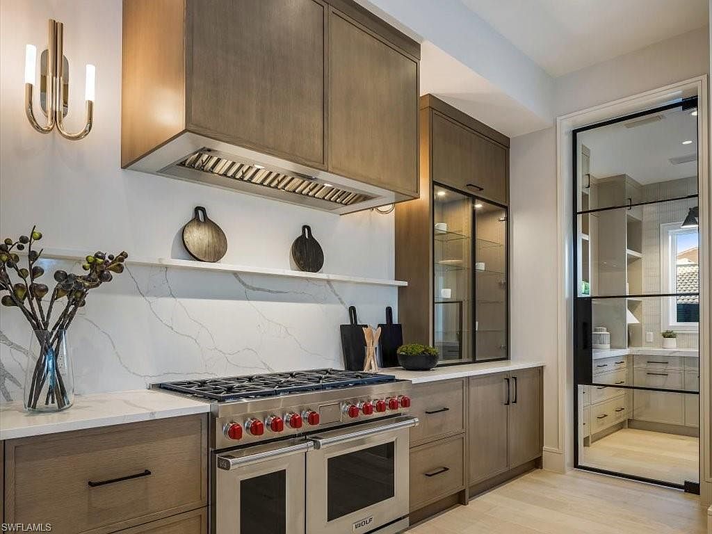 This is a well-lit kitchen featuring modern cabinetry with a light wood finish and sleek black hardware. A stainless steel range with red knobs is the focal point, complemented by a marble backsplash and countertop. A glass-paneled door offers a glimpse into another room, enhancing the sense of space and luxury.