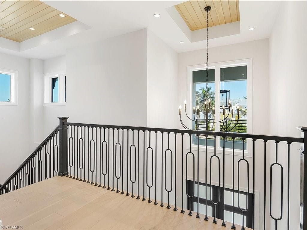 This interior shot showcases a modern hallway with a staircase. The black metal railing with unique oval designs adds a contemporary touch. Natural light floods the space through a large window, highlighting the light wood flooring and the elegant chandelier hanging from the ceiling.