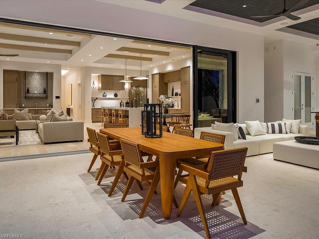 This interior shot showcases a dining area seamlessly connected to the living room and kitchen through a large sliding glass door. The dining area features a wooden table with woven chairs, while the living room offers comfortable seating with a white sofa and ottoman. The open floor plan and neutral color palette create a bright and inviting atmosphere, perfect for modern living.