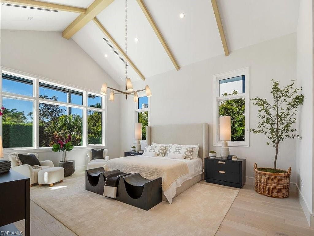 This is a bright and airy primary bedroom featuring a high vaulted ceiling with exposed beams and large windows that offer ample natural light and views of lush greenery. The room is furnished with a neutral-toned upholstered bed, dark wood nightstands, and a unique bench at the foot of the bed, all set on a light-colored area rug. The overall aesthetic is modern and luxurious, creating a serene and inviting space.