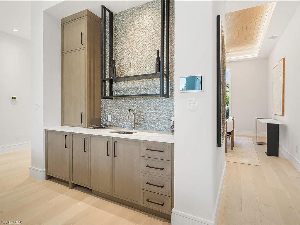 This interior shot showcases a modern kitchen area with light wood cabinetry, a white countertop, and a decorative backsplash. A built-in bar area features a metal frame with glass holders and a sink, adding a touch of sophistication. The space is well-lit and appears to be part of a larger, open-concept design, as suggested by the hallway visible on the right.