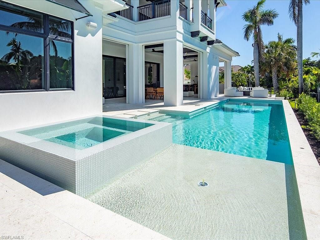 This image showcases a luxurious outdoor pool and spa area. The pool features clear turquoise water and is adjacent to a modern spa with white tile detailing. The surrounding architecture is contemporary with white columns and a balcony, complemented by palm trees and lush greenery, creating a serene and upscale atmosphere.