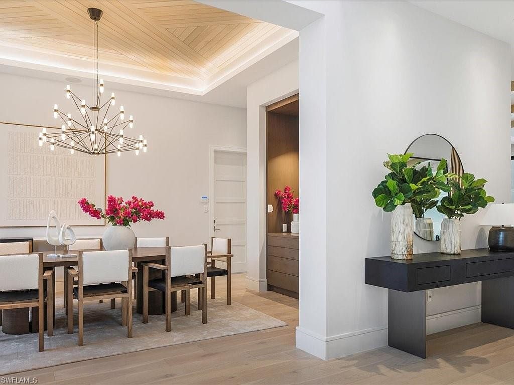 This interior shot showcases a modern dining room with a sophisticated aesthetic. The room features a wooden dining table surrounded by chairs with light-colored cushions, a striking chandelier, and a large piece of artwork on the wall. A console table with decorative plants and a round mirror adds to the room's elegance, while the hardwood floors and light walls create a bright and inviting atmosphere.