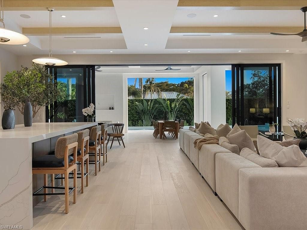 This is an interior shot of a modern living room that seamlessly blends indoor and outdoor spaces. The room features a large, comfortable sofa, a kitchen island with bar stools, and expansive sliding glass doors that open to a patio and garden area. The light wood flooring and neutral color palette create a bright and airy atmosphere, enhancing the sense of spaciousness and luxury.