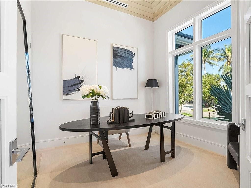 This interior shot showcases a modern office space featuring a dark, oval-shaped desk with unique leg supports, complemented by abstract artwork on the wall. A large window provides natural light and a view of lush greenery outside, enhancing the room's serene atmosphere. The space is decorated with a vase of white flowers, books, and decorative boxes, adding a touch of sophistication.