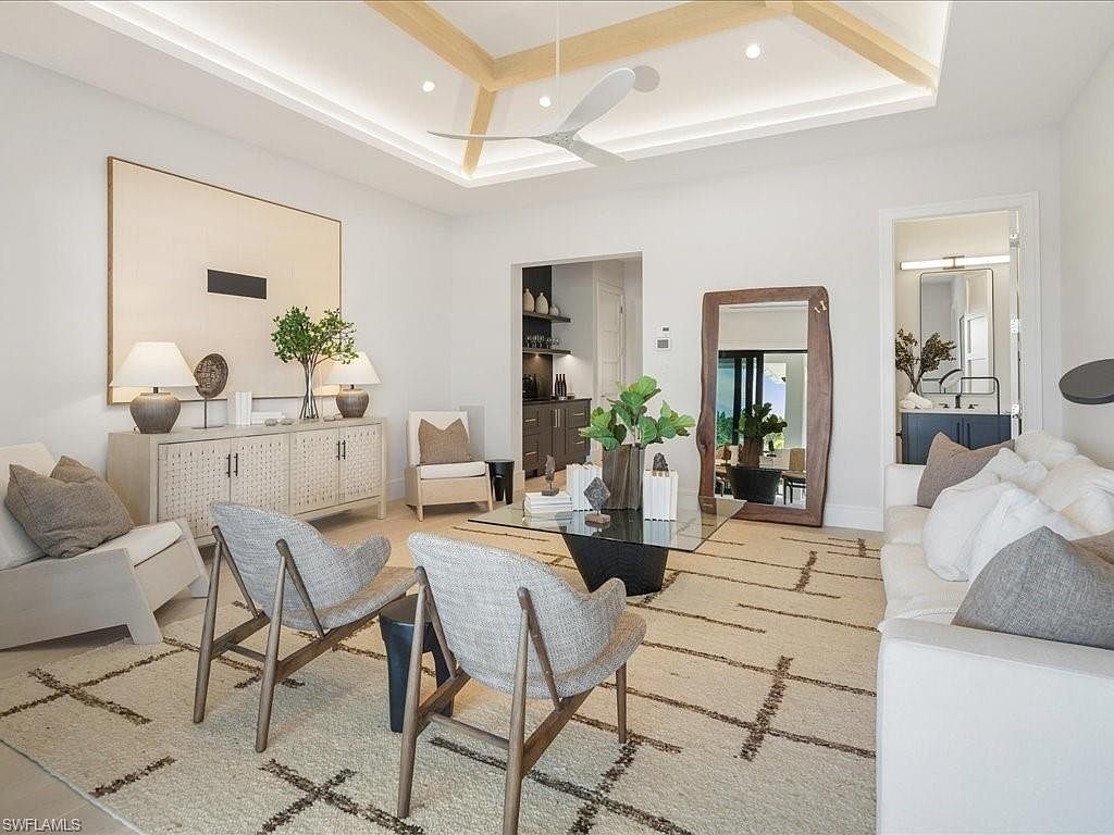 This is an interior shot of a modern living room. The room features a neutral color palette with white walls, light-colored furniture, and a patterned area rug. Key elements include a large piece of artwork, a wooden cabinet, two armchairs, a glass coffee table, and a white sofa, creating a bright and inviting space.